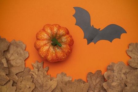 Autumn Halloween Composition with Pumpkin, Black Paper Bat, and Dry Leaves on Orange Backgroundの写真素材