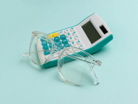 Clear Glasses Resting on Teal Calculator against Light Blue Backgroundの写真素材