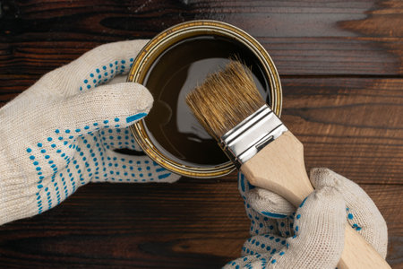 Hands in Protective Gloves Holding Paintbrush and Paint Can on Wooden Surface for DIY Projectの写真素材