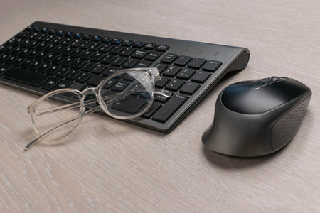 Modern Workspace with Wireless Keyboard, Mouse, and Reading Glasses on Wooden Deskの写真素材