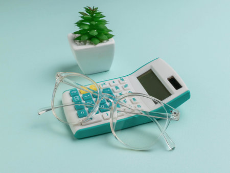 Clear Glasses Resting on Calculator Beside Small Succulent Plant on Light Blue Backgroundの写真素材