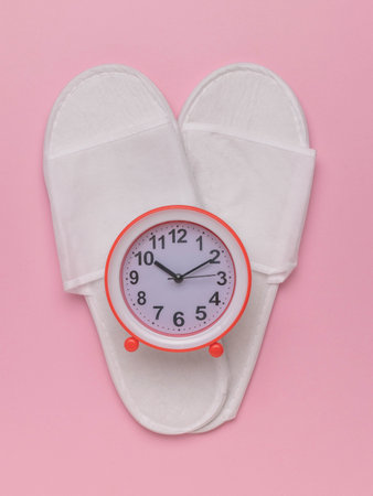 Cozy Morning Concept: White Slippers and Red Alarm Clock on Pale Pink Backgroundの写真素材