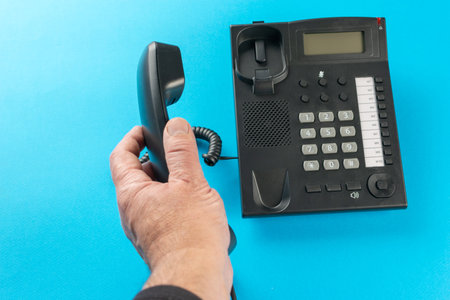 Hand Holding Landline Phone Handset Against Blue Background for Communication Conceptの写真素材