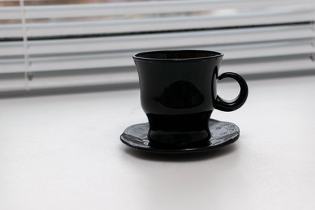 Sleek Black Coffee Cup on Saucer Near Window Blinds in Minimalist Settingの写真素材