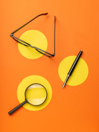 Modern Workspace: Black Magnifying Glass, Glasses, and Fountain Pen on Orange and Yellow Backgroundの写真素材
