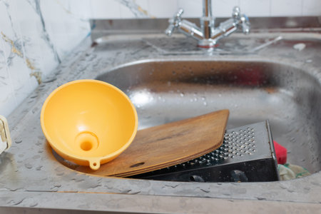 Messy Kitchen Sink with Cutting Board, Cheese Grater, and Yellow Funnel in Stainless Steel Basinの写真素材