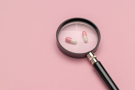 Magnifying Glass Over Capsules on Pink Background: Focus on Medicine and Healthcare Concepts in...