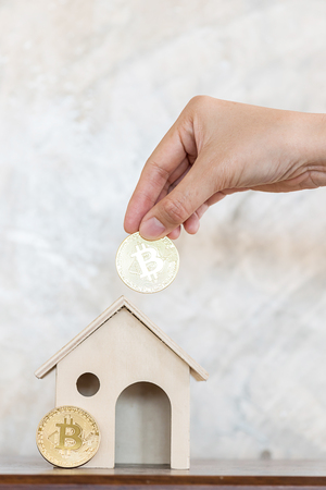 man hand put golden bitcoin into wood house modelの写真素材