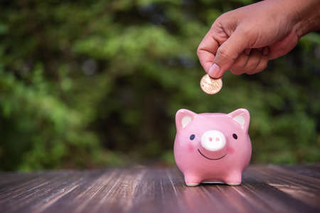 hand put a coin into piggy bank on wooden table with green natural leaves backgroundの写真素材