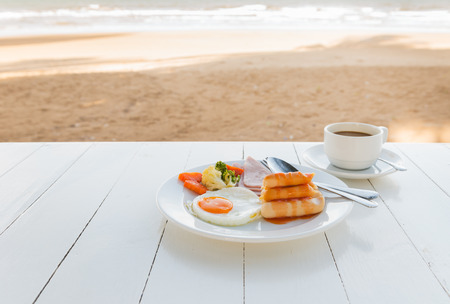 breakfast set and hot coffee on white table in morning and sand with sea  background.の写真素材