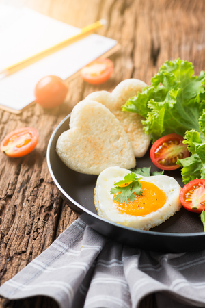 fried eggs and bread in the shape of a heart and fresh vegetables with blank notebook. Top view and wooden backgroundの写真素材