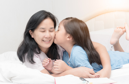 Sweet little girl is kissing her beautiful young mom in cheek while laying on bed at home, love and care concept.の写真素材