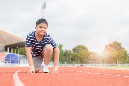fit and confident fat boy in starting position ready for running. kid athlete about to start a sprint looking at camera with bright sunlight. diet and exercise concept.の写真素材