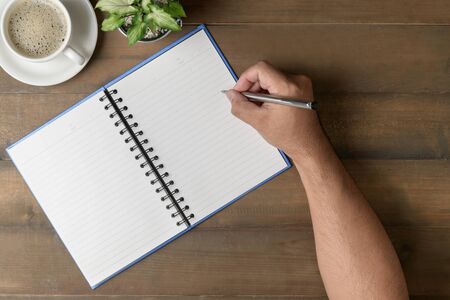Businessman writing on blank notebook with hot coffee on wood table, top view.