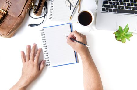 Business man write on blank notebook. White office desk table with laptop computer, cup of coffee...