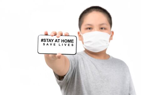 Child wear medical mask shows a mobile phone with stay at home save lives words isolated on whiteの写真素材