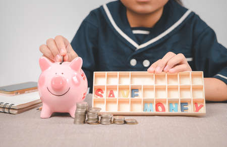Little girl  putting coin into piggy bank for saving with pile of coins on table at home and show save money text. A pink piggy bank are smile and happy while standing over pile of coins.の写真素材