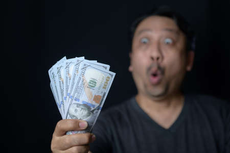 Asian handsome man holding bunch of dollars banknotes on black background scared and amazed with open mouth for surprise, disbelief faceの写真素材