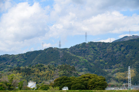 A scenic landscape featuring lush green mountains under a partly cloudy sky. Tall electricity transmission towers line the ridges, subtly blending with the natural surroundings.の写真素材