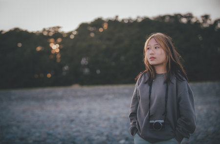 A young woman stands alone by a rocky shore, lost in thought as the setting sun filters through the...