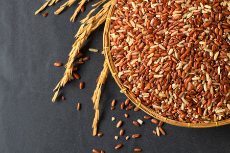 Close up of organic brown rice grains in a bamboo basket with golden rice stalks on black stone background. Perfect for concepts of agricultureの写真素材