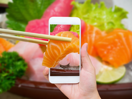 Female hand taking photo of sashimi sushi set with mobile phone and chopsticks hold salmon sashimi in mobile phoneの写真素材