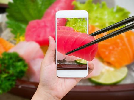 Female hand taking photo of sashimi sushi set with mobile phone and chopsticks hold tuna sashimi in mobile phoneの写真素材