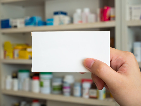 Close up pharmacist hand hold medicine box package with shelves of drugs in the pharmacyの写真素材