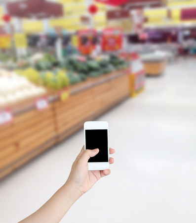Hand holding mobile phone with Vegetables and fruit on shelf in supermarket blurred backgroundの写真素材