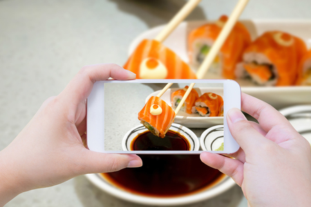 Female using mobile smart phone Taking photo of Salmon sushi in chopsticks dipping with soy sauceの写真素材
