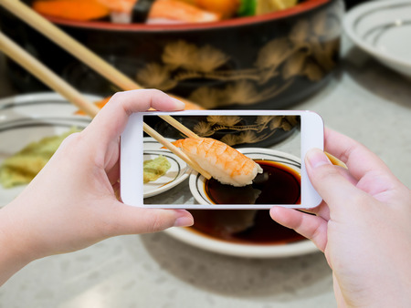 Female using mobile smart phone Taking photo of prawn sushi in chopsticks dipping with soy sauceの写真素材