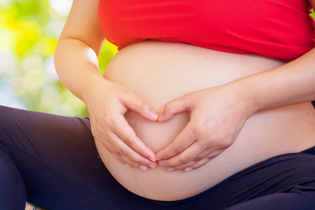 pregnant woman hands forming heart on her belly with nature background with bokeh light, pregnancy and love conceptの写真素材