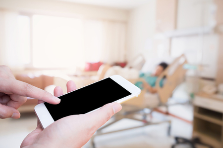 Female hand holding mobile smart phone with blurred hospital room with patient backgroundの写真素材