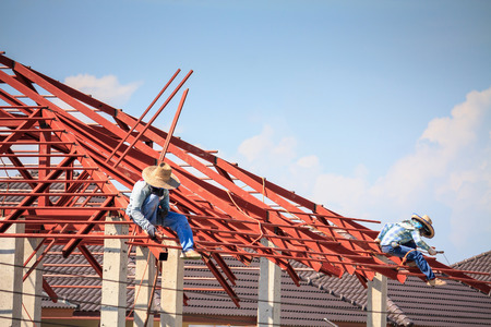Construction welder workers installing steel frame structure of the house roof at building construction site with clouds and skyの写真素材