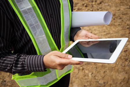 Asian business man construction manager and engineer worker in protective helmet hold blueprints paper on hand using tablet computer at building siteの写真素材