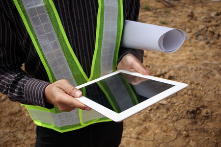 Asian business man construction manager and engineer worker in protective helmet hold blueprints paper on hand using tablet computer at building siteの写真素材