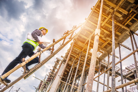 young professional engineer worker in protective helmet and blueprints paper on hand working on ladder at the house building construction siteの写真素材