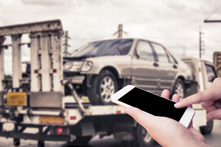 Hand using mobile smartphone for emergency roadside service with Broken car on tow truck after traffic accident backgroundの写真素材