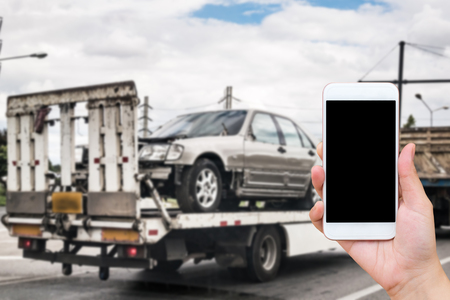 Hand using mobile smartphone for emergency roadside service with Broken car on tow truck after traffic accident backgroundの写真素材