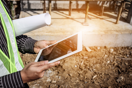 Asian business man construction manager and engineer worker in protective helmet hold blueprints paper on hand using tablet computer at building siteの写真素材