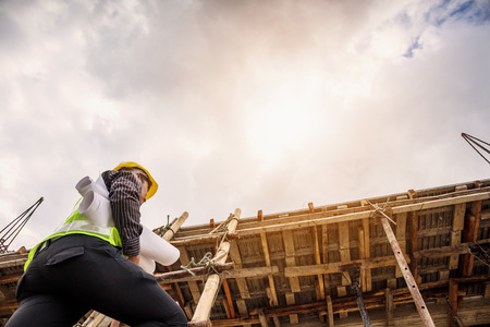 young professional engineer worker in protective helmet and blueprints paper on hand working on ladder at the house building construction siteの写真素材