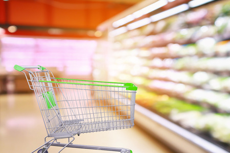 Supermarket aisle with fresh vegetables and fruit shelves interior blur background with empty shopping cart
