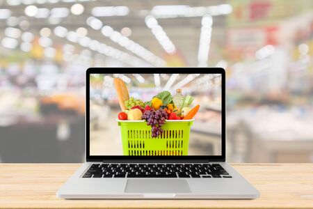 supermarket aisle blurred background with laptop computer and shopping basket on wood table grocery online conceptの写真素材