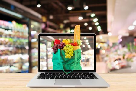 supermarket aisle blurred background with laptop computer and green shopping bag on wood table grocery online conceptの写真素材