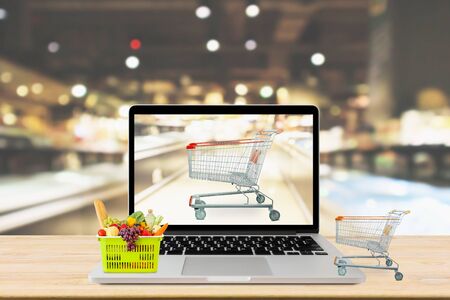 supermarket aisle blurred background with laptop computer and shopping cart on wood table grocery online conceptの写真素材