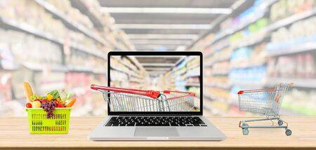 supermarket aisle blurred background with laptop computer and shopping cart on wood table grocery online conceptの写真素材