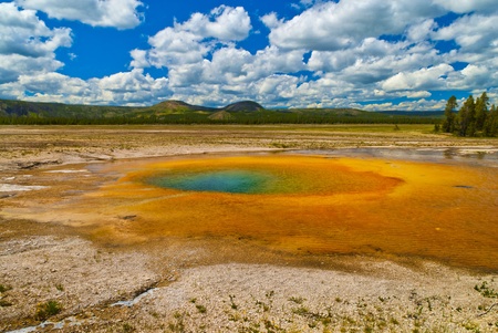 Beautiful cerulean geyser surrounded by colorful layers of bacteria, against cloudy blue sky.の写真素材