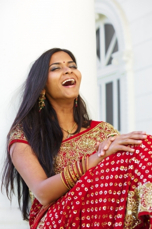 A young, 20 something, Indian woman with long black hair, dressed in traditional Indian attire (saree), is laughing as she rests against a tall white column on a bright sunny day の写真素材