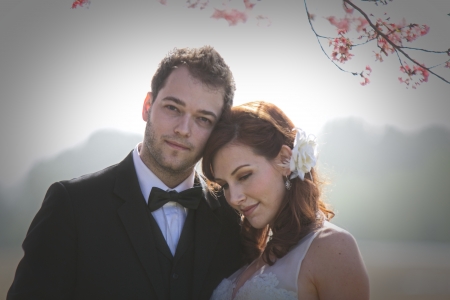 Moody backlighting illuminates a young, 20 something, man dressed in a black suit with a bow tie and woman in a sleeveless white wedding gown, standing next to each other.の写真素材
