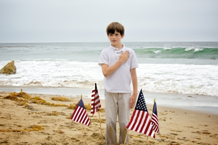 A young boy, with sand all over his hands and pants, plants four American flags into the sand at the beach then stands among them with his hand to his heartの写真素材